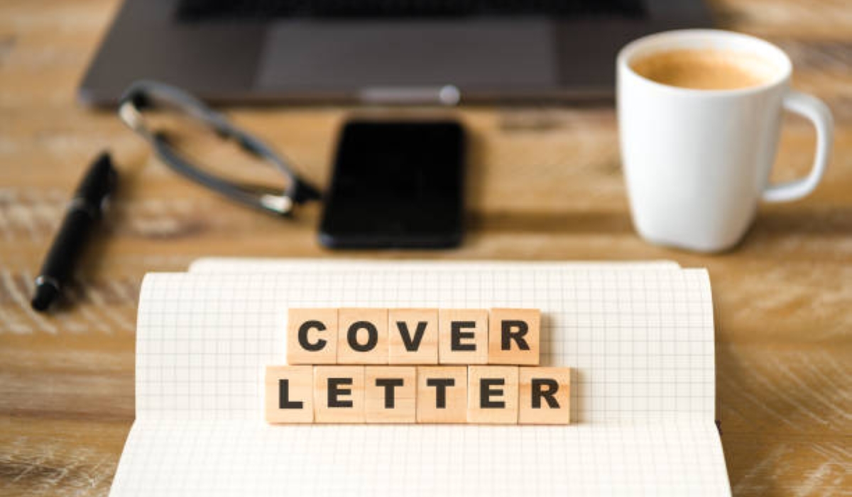 Closeup on notebook over wood table background, focus on wooden blocks with letters making COVER LETTER words. Business concept image. Laptop, glasses, pen and mobile phone in a defocused background.