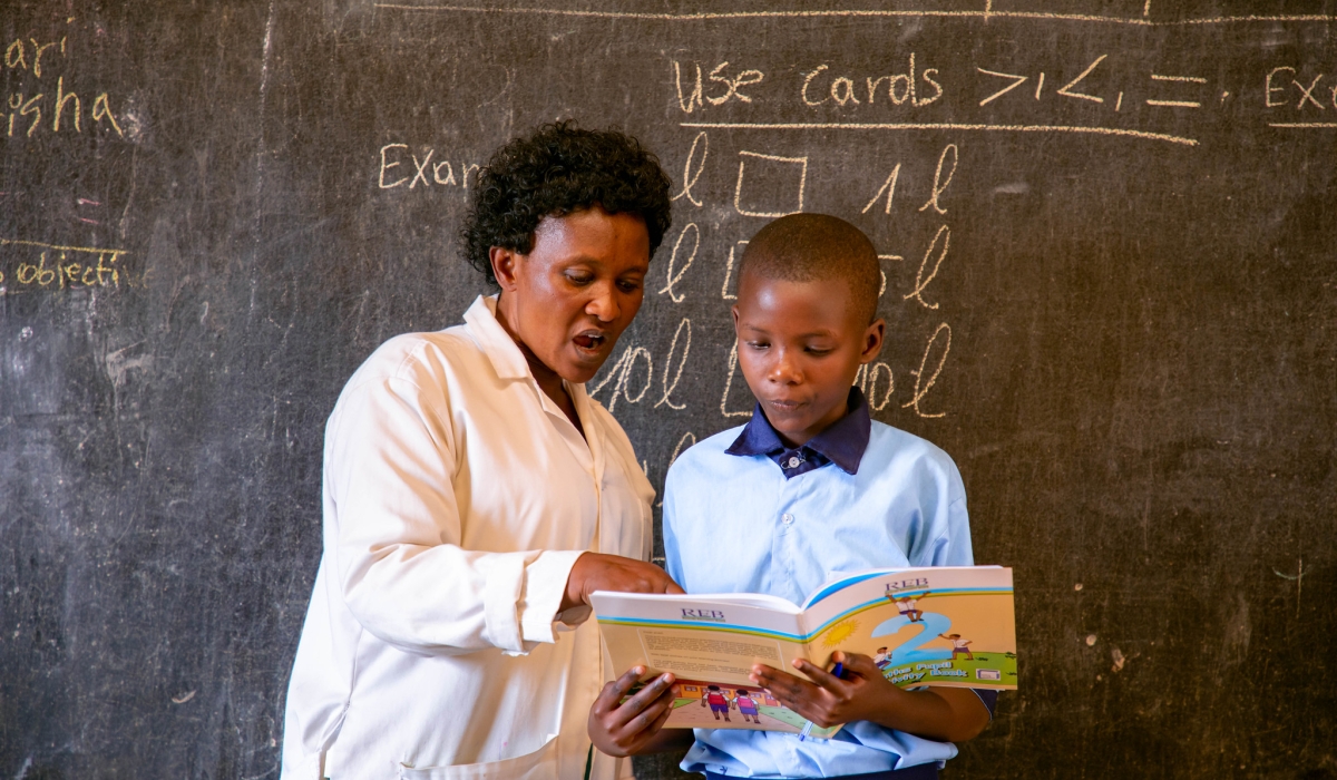 A teacher helps a child while reading a book at Ecole Primaire Kimisange. REB has revealed that Rwanda needs Rwf14 billion to ensure that every pupil or student gets a textbook at school. Photo by Dan Gatsinzi