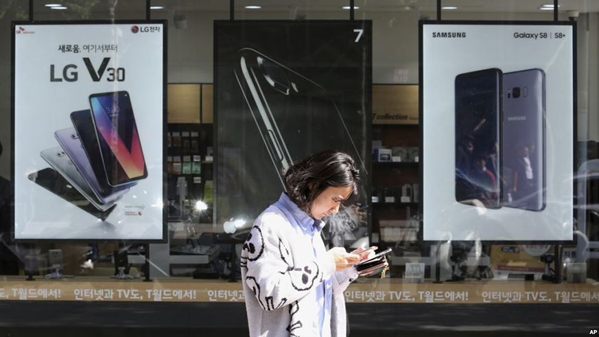 A woman checks her phone as she walks by posters adverting smartphones at a mobile phone store in Seoul, South Korea. Net photo.