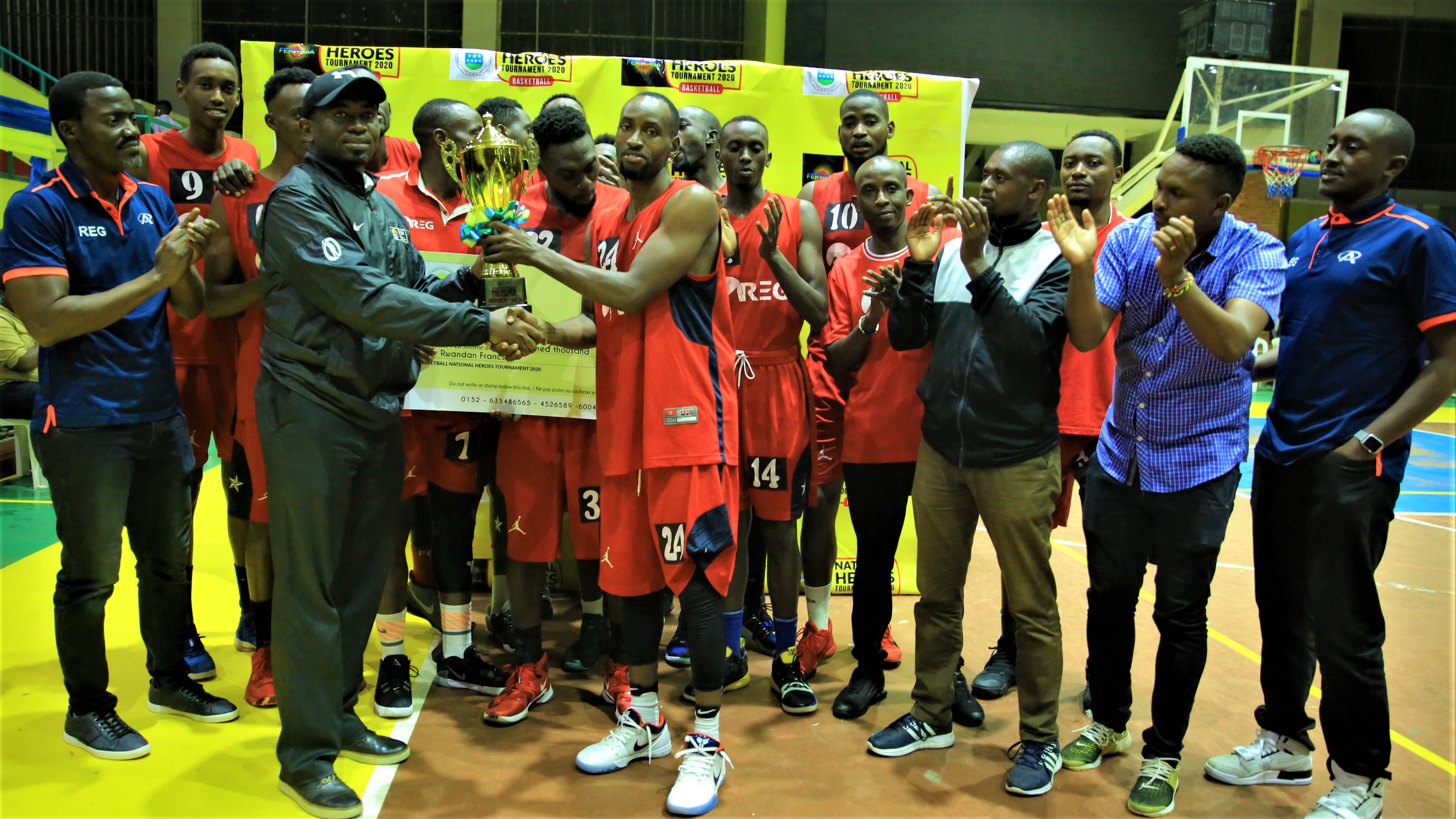 Didier Shema Maboko, Permanent Secretary of the Ministry for Sports, hands over the trophy to REG captain Ali Kubwimana Kazingufu as teammates and coaching staff in the background celebrated the milestone after beating APR 71-62 to retain the Heroes Cup for a third consecutive time at Amahoro Stadium on Sunday night. Photo: 